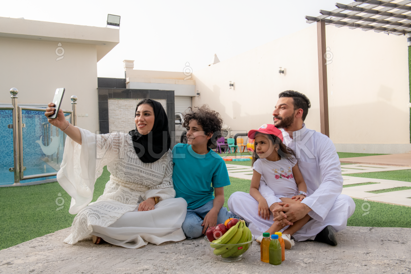  امرأة عربية سعودية محجبة تجلس مع عائلتها في حديقة المنزل بينما تحمل الجوال لإلتقاط صورة تجمعهم معاً  