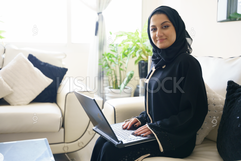  امرأة عربية محجبة ترتدي عباية وتنظر لليسار وتبتسم بينما تجلس على الكنب وتستخدم اللابتوب  