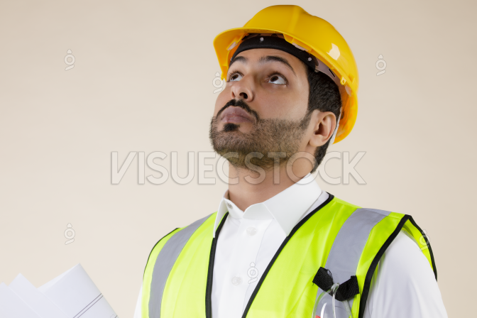  صورة مهندس سعودي يرتدي سترة وخوذة أمان يحمل قائمة بمشروع بناء أثناء النظر أعلاه  