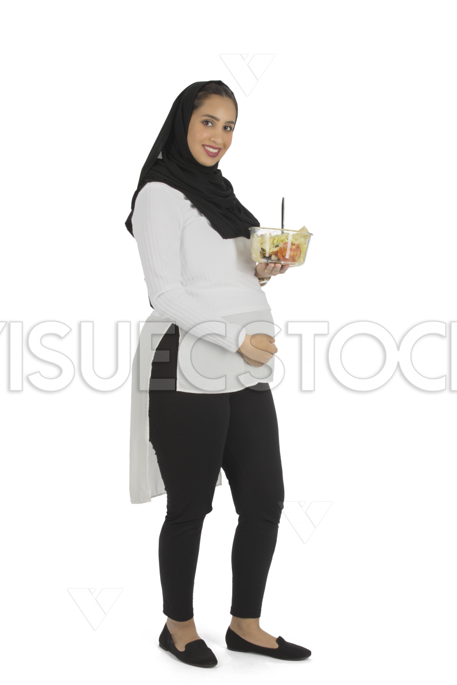  إمأة عربية سعودية حامل تقف مبتسمة وتمل في يدها طبق خضوات وتضع يدها عل بطنه  