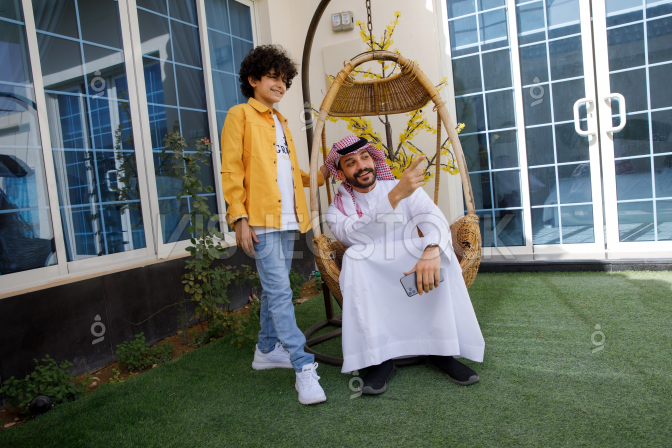  رجل عربي سعودي يرتدي ثوب أبيض وشماغ وعقال ويجلس على أرجوحة منزلية في حديقة المنزل وإبنه يقف بجواره يبتسمون وينظرون حيث يشير الأب.  طفل عربي سعودي يرتدي بنطلون جينز وقميص يقف بجوار والده في حديقة المنزل وينظر حيث يشير والده بيده اليمين .  أب عربي سعودي يستمتع بقضاء وقت مع إبنه في حديقة المنزل بينما يجلس على أرجوحة منزلية  
