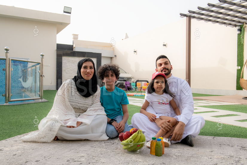  إمرأة عربية خليجية محجبة تجلس بجوار أفراد أسرتها في حديقة المنزل وتبتسم بينما ينظرون إلى اليمين  