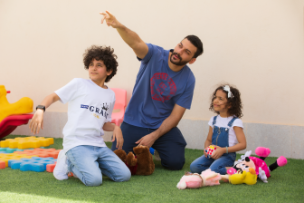 رجل عربي سعودي يجلس في حديقة المنزل مع أبنائه أثناء اللعب ويشير بيده اليمين للأعلى