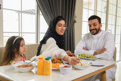 امرأة عربية سعودية محجبة تبتسم وتقدم الطعام لأسرتها بينما يستمتعون بتناول الطعام معاً في المنزل