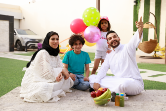 عائلة عربية سعودية تستمتع بالجلوس في حديقة المنزل وينظرون للأعلى حيث يشير الأب بيده ويبتسم.  رجل عربي سعودي يجلس مع عائلته في الحديقة ويبتسم وينظر لأعلى اليمين.  طفلة عربية سعودية تقف خلف عائلتها وتحمل بالونات بينما تستمتع العائلة بالهواء الطلق في حديقة المنزل