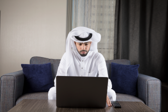 رجل عربي سعودي يجلس على كنة يرتدي ثوب أبيض غترة ويعمل من المزل على اللابتوب