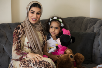 امرأة عربية محجبة تجلس على أريكة بالمنزل وتبتسم وبجوارها تجلس إبنتها مع العابها