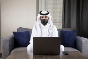 رجل عربي سعودي يجلس عل كنبة يرتدي كمامة سوداء ونظر إلى الأمام، رجل أعمال عبي خليي يعمل من امنزل