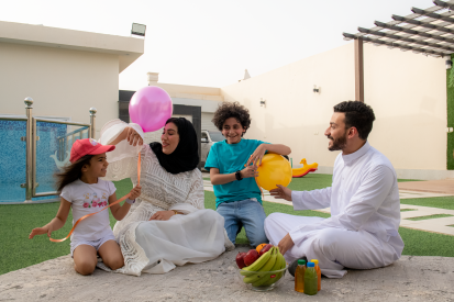 عائلة عربية سعودية تستمتع بقضاء وقت في حديقة المنزل . امرأة عربية سعودية ترتدي عباية بيضاء وحجاب أسود وتنظر لليسار وتبتسم وبجانبها طفلتها تشاركها اللعب بالبالونات