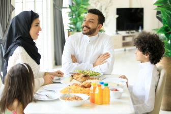 عائل عربية خليجية ستمتع بتناول الطام معاً في المنزل ،  رجل عربي خليجي يبسم وينظر لزوجته أثناء الجلوس مع أطالهما على طاولة لطعام بالمنزل