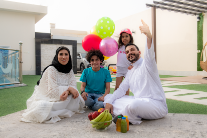 أسرة عربية سعودية سعيدة في حديقة داخلية تبتسم بينما تنظر إلى الكاميرا ويشير الأب للأعلي