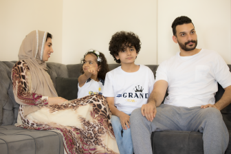 أسرة عربية سعودية تجلس معًا على أريكة في المنزل وتستمتع بمشاهدة التلفاز بينما تشير الابنة إلى شيء أمامها