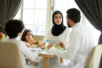 عائلة عربية خليجية تسمتع بتاول الطعا معاً،  مرأة عربة خليجية محجبة تبتسم بينما تجلس مع أفراد أسرتها على اولة الطعم في المنزل