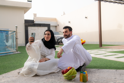 جل عربي سعودي يرتدي ثوب أبيض ويجلس بجوار زوجته ي حدقة المنزل .  ارأة عربية سعودية ترتي عباية وحجاب تجلس مع زوجها في ديقة المنزل وتلتط هما صورة بالجوال. عائلة عربية سعودة تستمتع بالجلوس في حديقة المنزل
