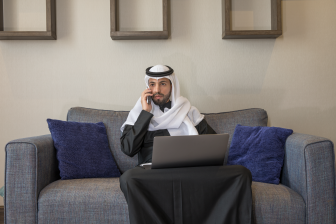 رجل عربي سعودي يجلس على الكنبة يرتدي ثوب رمادي و غترة بيضاء يعمل من المنزل على اللابتوب ويتحدث على الجوال