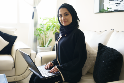 امرأة عربية محجبة تنظر لليار مع الابتسامة بينما تستخدم اللابتوب وهي جلس على أريكة بالمنزل