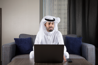 رجل أعمال عربي سعودي مبتسم يجلس على كنبة يرتدي ثوب أبيض وغترة وينظر إلى يساره