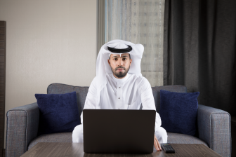 رجل عربي سعوي جلس على كنبة يتدي ثوب أبيض وغترة وأمامه لابتوب