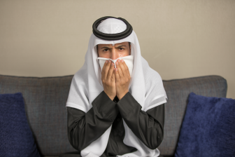 رجل عربي سعودي يرتدي ثوب رمادي و غترة بيضاء، يجلس على كنبة، يعطس او يسعل في منديل