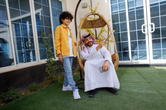رجل عربي سعودي يرتدي ثوب أبيض وشماغ وعقال ويجلس على أرجوحة منزلية في حديقة المنزل وإبنه يقف بجواره يبتسمون وينظرون حيث يشير الأب.  طفل عربي سعودي يرتدي بنطلون جينز وقميص يقف بجوار والده في حديقة المنزل وينظر حيث يشير والده بيده اليمين .  أب عربي سعودي يستمتع بقضاء وقت مع إبنه في حديقة المنزل بينما يجلس على أرجوحة منزلية
