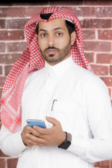 صورة لرجل سعودي يرتدي ثوًا تقيديًا يقف ويحمل هاتفًا محمولًا أثناء النظر إلى الكايرا