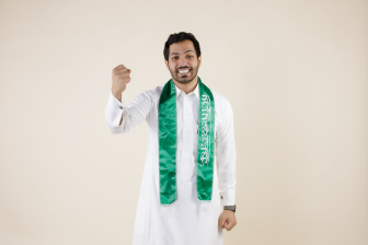رجل سعودي يلف علم السعوديه حول عنقه ويبتسم بحماس ويرفع قبضة يديه