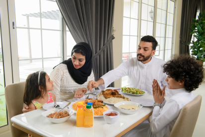مرأة عربية سعودي محجبة تبتسم وتظر لإبنتها أثناء اللوس مع أسرتها لى اولة الطعام. جل عبي سعودي يرتدي ثوب أبيض يتناو الطعم مع أسرته ي المنل