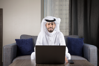 رجل عربي سعودي مبتسم يجلس على كنبة يرتدي ثوب أبيض وغترة وأمامه لابتوب