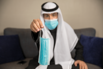 رجل عربي سعودي يرتدي قناع وجه (كمامة) بينما يمسك قناع آخر في يده اليمين