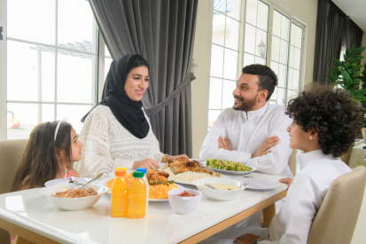 عائلة عربية سعودية تجلس معاً على طاولة الطعام في المنزل ويبتسمون.. رجل عربي سعودي ينعم بقضاء وقت ممتع مع أسرته ويستمتعون بتناول الطعام في المنزل