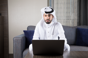 رجل عربي سعودي يجلس على كنبة يرتدي ثوب أبيض وغترة يستخدم اللابتوب وينظر إلى الأمام