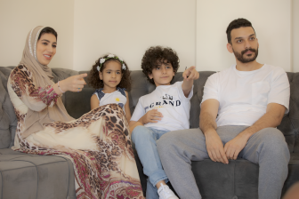 عائلة عربية سعودبة تستمتع بالجلوس معاَ في المنزل ومشاهدة التليفزيون  امرأة عربية سعودية محجبة تشير بيدها لليمين أثناء الاستمتاع بمشاعدة التلفاز مع أفراد أسرتها