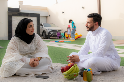 رجل عربي سعودي يرتدي ثوب أبيض يستمتع بالجلوس مع أسرته في حديقة المنزل وبجواره زوجته بينما يلعب أطفالهما في الخلف